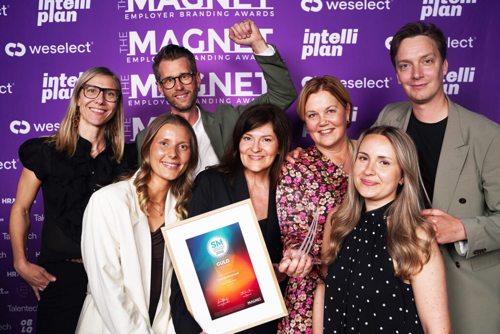 Ett team på sju personer poserar glatt framför en lila pressvägg med logotyperna "The Magnet Employer Branding Awards", Weselect och Intelliplan. Två personer i mitten håller upp ett inramat SM Guld-diplom och en glastrofé från Magnet 2025. En person i bakgrunden håller upp en knytnäve i jubel.